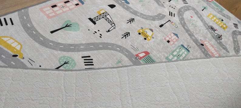 May include: A gray and white play mat with a city theme. The mat features a winding road, buildings, trees, cars, and a construction crane.