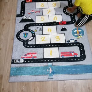 May include: A grey and blue play mat with a road and airport design. The mat features numbers 1-10, a traffic light, and various toy vehicles including a fire engine, ambulance, police car, and helicopter.