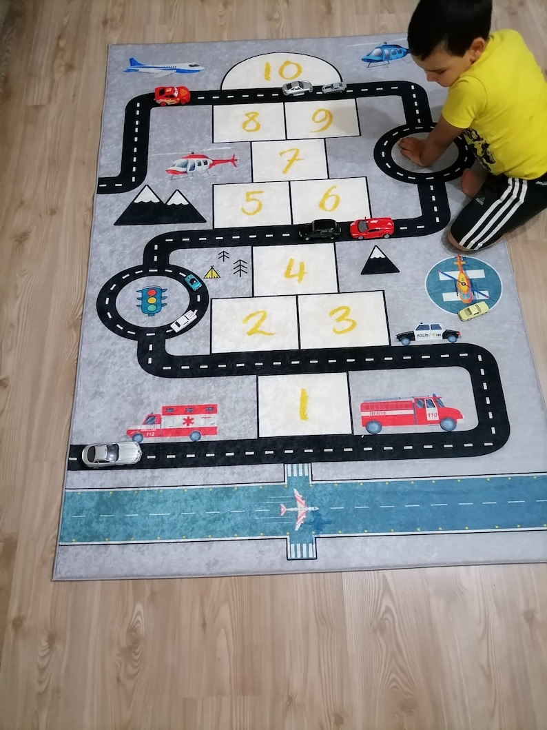May include: A grey and blue play mat with a road and airport design. The mat features numbers 1-10, a traffic light, and various toy vehicles including a fire engine, ambulance, police car, and helicopter.