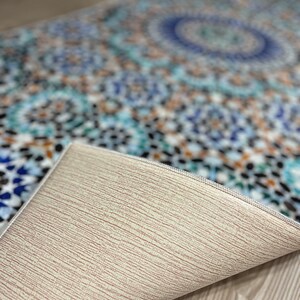 K&ouml;nnte beinhalten: Ein gemusterter Teppich mit einem Mosaikdesign in Blau-, T&uuml;rkis- und Braunt&ouml;nen. Die Unterseite des Teppichs ist hellbeige und zeigt seine Textur. Das Design zeigt geometrische Formen und ein zentrales kreisf&ouml;rmiges Motiv.