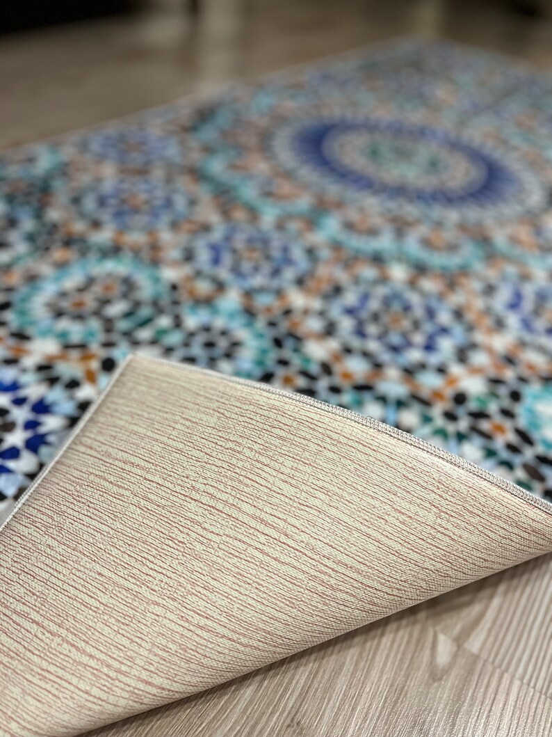 K&ouml;nnte beinhalten: Ein gemusterter Teppich mit einem Mosaikdesign in Blau-, T&uuml;rkis- und Braunt&ouml;nen. Die Unterseite des Teppichs ist hellbeige und zeigt seine Textur. Das Design zeigt geometrische Formen und ein zentrales kreisf&ouml;rmiges Motiv.