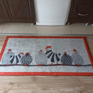 May include: A rectangular kitchen mat with a design of chickens in black, white, and grey. The chickens are on a dark grey line against a silver background, with a red border. The mat is placed on a wooden floor, likely in a kitchen.