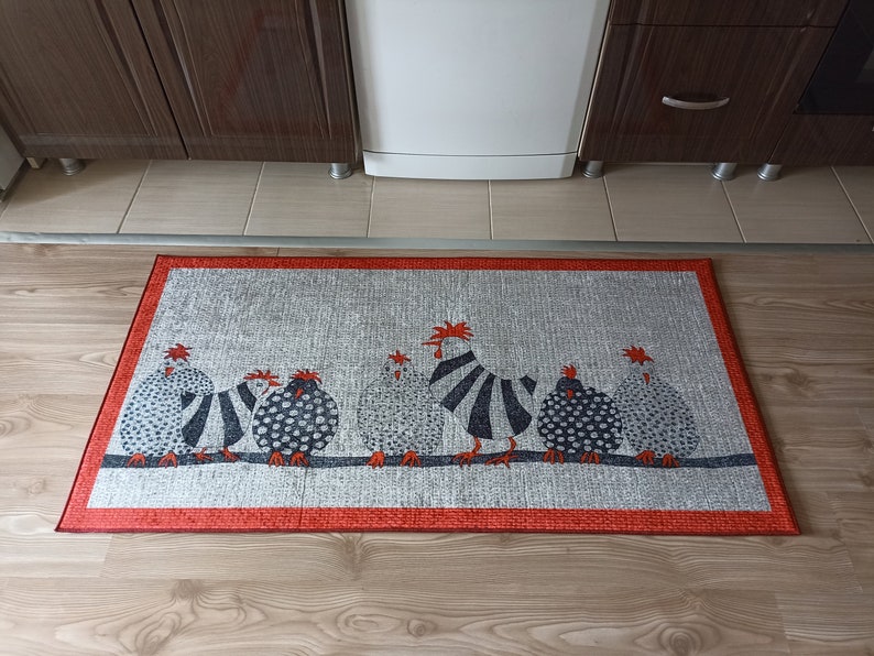 May include: A rectangular kitchen mat with a design of chickens in black, white, and grey. The chickens are on a dark grey line against a silver background, with a red border. The mat is placed on a wooden floor, likely in a kitchen.