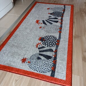 May include: Rectangular grey rug with a red border. The rug features a repeating pattern of stylised chickens in black, white, and blue. The chickens are perched on a dark brown branch, with red accents.