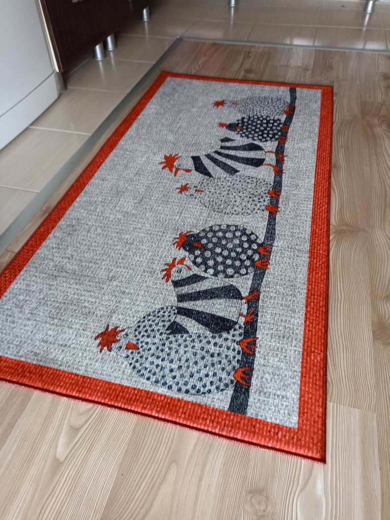 May include: Rectangular grey rug with a red border. The rug features a repeating pattern of stylised chickens in black, white, and blue. The chickens are perched on a dark brown branch, with red accents.