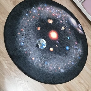Puede incluir: Una alfombra redonda que presenta una detallada representaci&oacute;n del sistema solar sobre un fondo oscuro y estrellado. Los planetas, las estrellas y las galaxias est&aacute;n ilustrados en colores vibrantes, creando un tema c&oacute;smico. La alfombra est&aacute; dise&ntilde;ada para colocarse en el suelo.