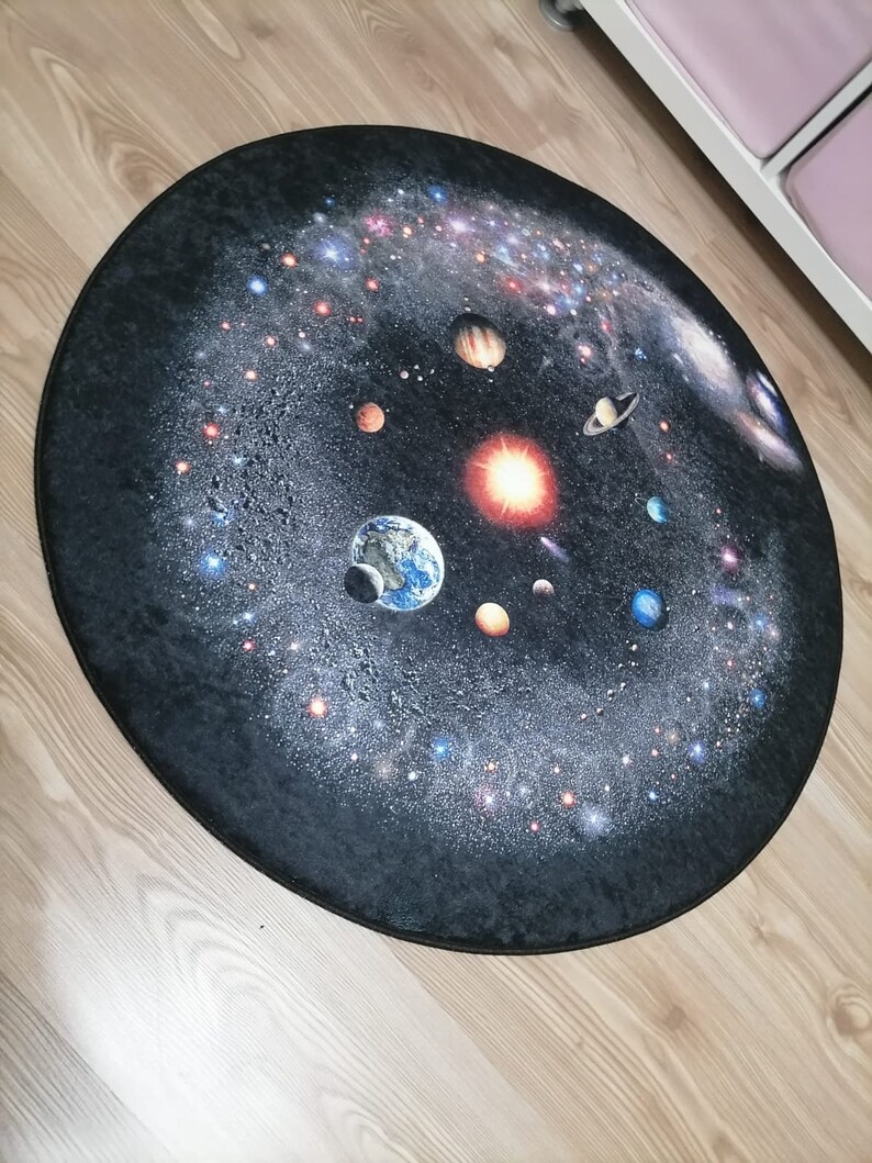 Puede incluir: Una alfombra redonda que presenta una detallada representaci&oacute;n del sistema solar sobre un fondo oscuro y estrellado. Los planetas, las estrellas y las galaxias est&aacute;n ilustrados en colores vibrantes, creando un tema c&oacute;smico. La alfombra est&aacute; dise&ntilde;ada para colocarse en el suelo.