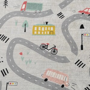 May include: A gray and white play mat with a winding road design. The road has cars, a bus, a bicycle, a crane, and a building. There are trees and flowers along the road.