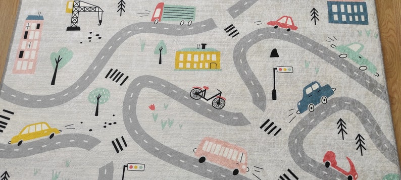 May include: A gray and white play mat with a winding road design. The road has cars, a bus, a bicycle, a crane, and a building. There are trees and flowers along the road.