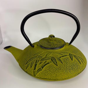 May include: A lime green cast iron teapot featuring a textured surface with a raised bamboo leaf pattern. The teapot has a black arched handle, a matching circular lid with a small knob, and a short spout extending from the side of the body.