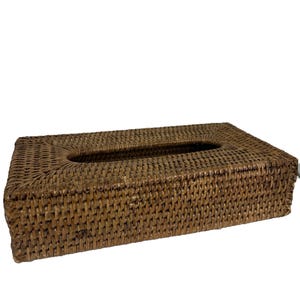 May include: A rectangular tissue box cover crafted from dark brown woven rattan. The item features a tight, uniform weave pattern throughout its exterior surfaces and includes a centered, elongated oval opening on the top for dispensing tissues.