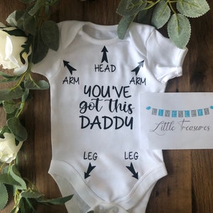 May include: White baby grow with black text that reads "You've got this Daddy" with arrows pointing to the head, arms, and legs. The grow is on a wooden surface with white roses and green leaves.