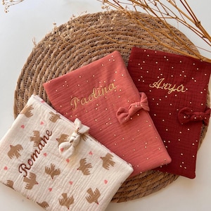 Peut inclure: Trois pochettes rectangulaires en tissu avec des nœuds. L'une est crème avec des motifs d'oiseaux marron et de cœurs roses et le nom Romane. Une est rose poudré avec des points dorés et le nom Paolina. Une est rouge foncé avec des points dorés et le nom Arya.
