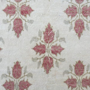 May include: A close-up of a white rug with a repeating pattern of pink floral designs. The rug has a subtle texture and a worn look.