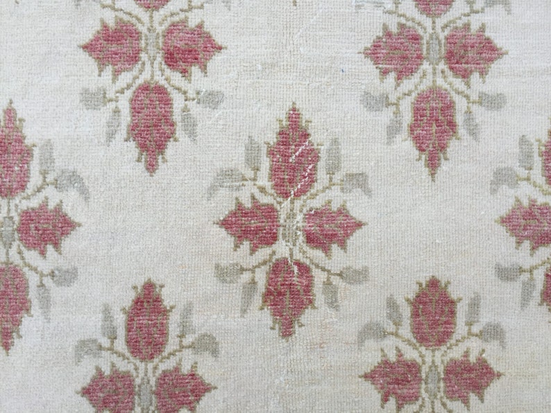May include: A close-up of a white rug with a repeating pattern of pink floral designs. The rug has a subtle texture and a worn look.