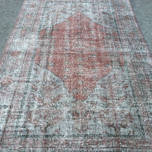 Op de afbeelding: Een vintage tapijt met een vervaagd rood en wit patroon. Het tapijt heeft een ruitvormig ontwerp in het midden en een rand rond de randen.