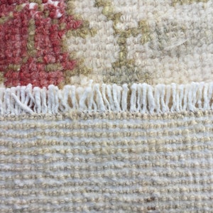 May include: Close-up of a white and beige woven rug with a fringe border. The rug has a subtle pattern of small squares.