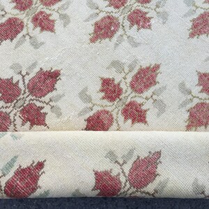 May include: A cream-colored rug with a repeating pattern of red and green floral motifs. The rug is rolled up, showing the underside of the fabric.
