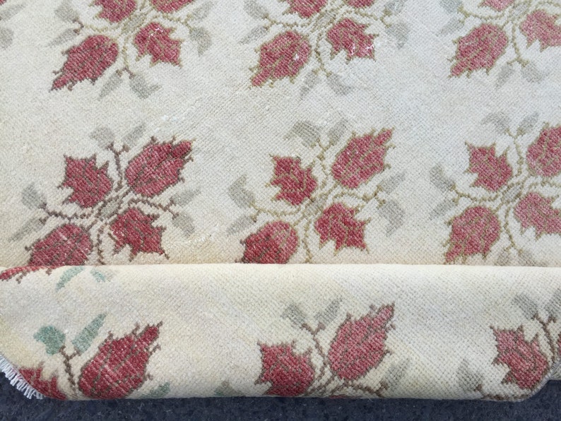 May include: A cream-colored rug with a repeating pattern of red and green floral motifs. The rug is rolled up, showing the underside of the fabric.