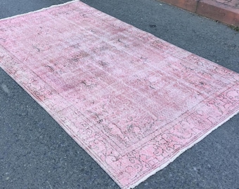 May include: A rectangular area rug featuring a faded pink base color with intricate, distressed dark gray floral and geometric patterns throughout the center and a wide, detailed border framing the entire piece with fringed edges.