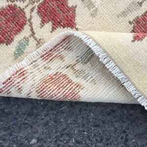 May include: Close-up of a cream-colored rug with a floral pattern. The rug has a fringe border and is folded in half. The rug is lying on a dark gray surface.