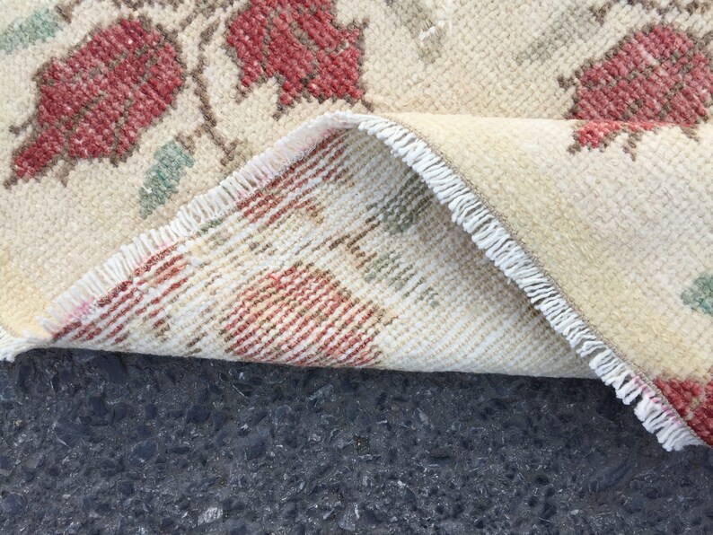 May include: Close-up of a cream-colored rug with a floral pattern. The rug has a fringe border and is folded in half. The rug is lying on a dark gray surface.