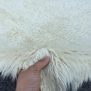 Peut inclure: Gros plan sur un tapis crème et touffu. Le tapis a de longues fibres douces et un aspect texturé. Les fibres sont tenues par une main, montrant la souplesse et l'épaisseur du tapis. Le tapis semble être fait de matériaux naturels.