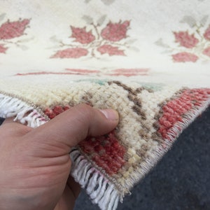 May include: Close-up of a white rug with a red floral pattern. The rug has a fringe border and is being held by a hand.