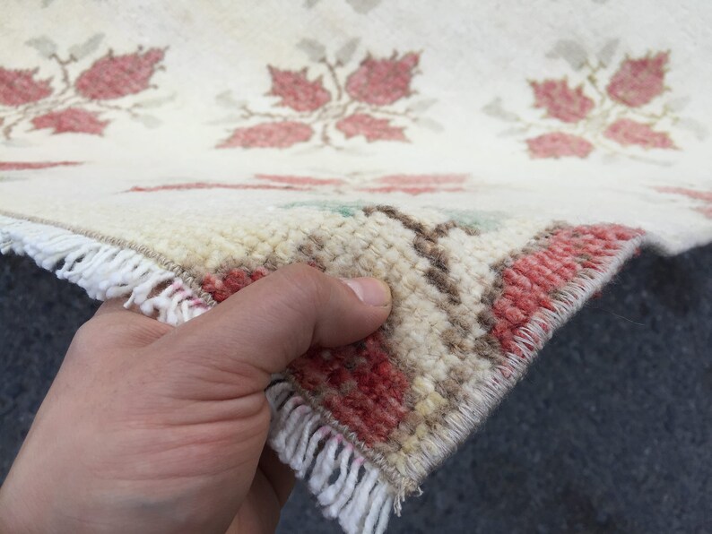 May include: Close-up of a white rug with a red floral pattern. The rug has a fringe border and is being held by a hand.