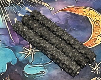 May include: Three cylindrical black candles featuring a textured honeycomb pattern on the exterior. Each candle has a visible white wick at the top end. The candles are arranged in a parallel row, showcasing the detailed hexagonal surface texture.