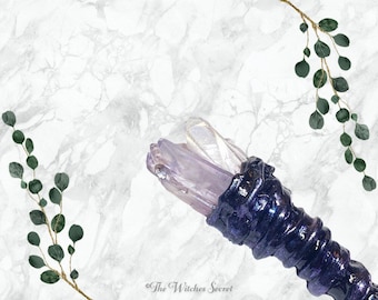 Puede incluir: Una varita mágica artesanal con un diseño de cristal morado y transparente. La varita presenta un mango texturizado, de color púrpura oscuro y está rematada con dos cristales. El fondo es un patrón de mármol con follaje verde.