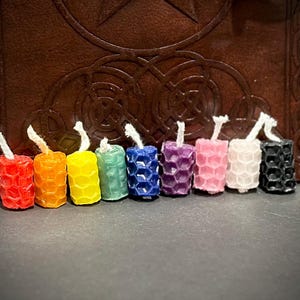 May include: A row of nine small cylindrical candles featuring a textured honeycomb pattern. The candles are arranged in a spectrum of colors including red, orange, yellow, green, blue, purple, pink, white, and black, each with a white cotton wick.