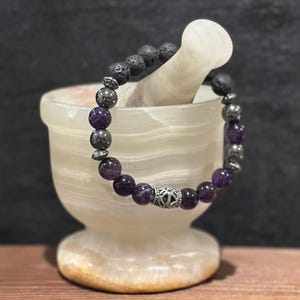 May include: A beaded bracelet featuring amethyst, lava stone, and silver-toned beads. The bracelet is displayed on a white mortar and pestle. The amethyst beads are a deep purple color.