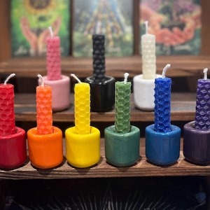 May include: A collection of nine cylindrical candles with a honeycomb texture, each set in a matching coloured ceramic holder. The candles and holders are arranged in a spectrum of colours including red, orange, yellow, green, blue, purple, pink, black, and white.