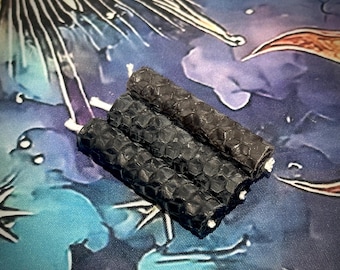 May include: Three cylindrical black candles with a textured honeycomb pattern on the exterior. Each candle features a visible white cotton wick at the top and bottom, arranged in a parallel cluster to form a single unit of three connected pillars.
