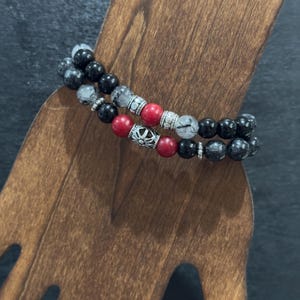 May include: Two beaded bracelets with black, red, and clear beads. The bracelets are displayed on a wooden stand. The beads are round and strung together with silver-coloured spacers. The bracelets have a mix of textures and colours.