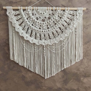 以下が含まれることがあります： 円形のデザインとフリンジが付いた白いマクラメの壁掛け。コットンコードで作られており、木製のダボに付けられています。