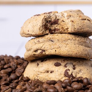 May include: A stack of three chocolate chip cookies with a bite taken out of the top cookie. The cookies are sitting on a bed of coffee beans.