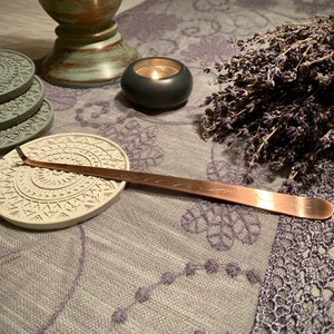 May include: A set of four gray and white patterned coasters with a copper-colored incense stick holder on top of one of the coasters. A lit candle and a bunch of dried lavender are also in the image.