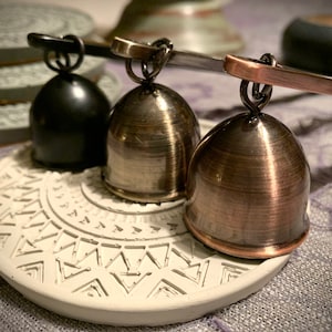 May include: Three decorative bells with a silver, copper, and black finish. The bells are hanging from a metal rod and are sitting on a white ceramic coaster with a geometric pattern.