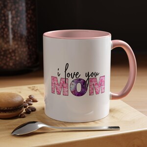 Może przedstawiać: Biały ceramiczny kubek z różowym wnętrzem i uchwytem. Kubek zawiera napis "i love you MOM" czarnym pismem oraz różowo-fioletowy kwiatowy wzór. Na drewnianej powierzchni leżą łyżka i ciastko.
