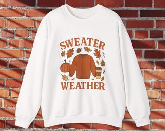 May include: White sweatshirt with a fall-themed graphic. The design features a brown sweater, pumpkin, coffee cup, and autumn leaves, with the words "SWEATER WEATHER" in brown lettering.