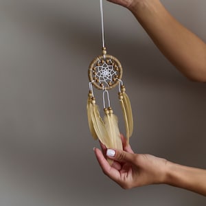 Op de afbeelding: Een kleine droomvanger met bruine en witte kralen en beige veren die aan een wit touwtje hangen.