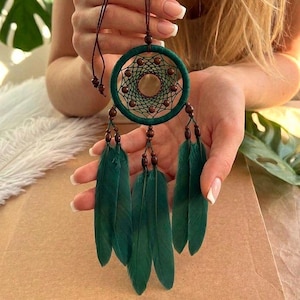 May include: A handcrafted dreamcatcher with a teal green hoop, intricate web design, and dark green feathers. The dreamcatcher features wooden beads and a brown cord for hanging. The design is held by a person.