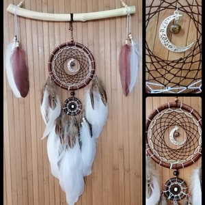 May include: A brown and white dreamcatcher with feathers hanging from the bottom. The dreamcatcher has a crescent moon charm with a brown stone in the center. The dreamcatcher is hanging from a wooden branch.