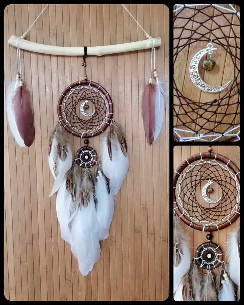 May include: A brown and white dreamcatcher with feathers hanging from the bottom. The dreamcatcher has a crescent moon charm with a brown stone in the center. The dreamcatcher is hanging from a wooden branch.