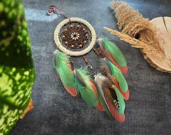May include: A handcrafted dreamcatcher with a beige woven hoop and a central starburst design in brown. Green and orange feathers hang below, accented with small beads. The dreamcatcher is suspended by a brown cord.