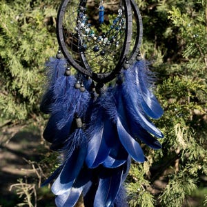 Puede incluir: Un atrapasue&ntilde;os artesanal con marco negro y plumas azul marino. Cuenta con una red de hilo negro, cuentas y un colgante de cristal azul. Las plumas est&aacute;n dispuestas en un patr&oacute;n en capas, creando un dise&ntilde;o visualmente atractivo.