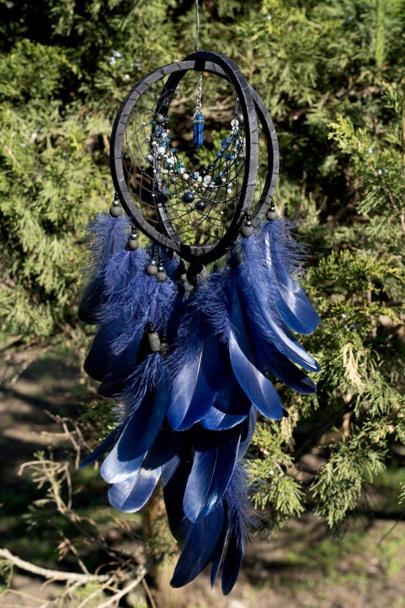 Puede incluir: Un atrapasue&ntilde;os artesanal con marco negro y plumas azul marino. Cuenta con una red de hilo negro, cuentas y un colgante de cristal azul. Las plumas est&aacute;n dispuestas en un patr&oacute;n en capas, creando un dise&ntilde;o visualmente atractivo.