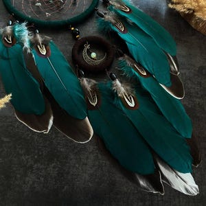 May include: A teal dreamcatcher with large, vibrant teal feathers, accented with brown and white tips. A circular pendant with a crescent moon and a small, light-colored stone hangs in the center. The dreamcatcher is a decorative item.
